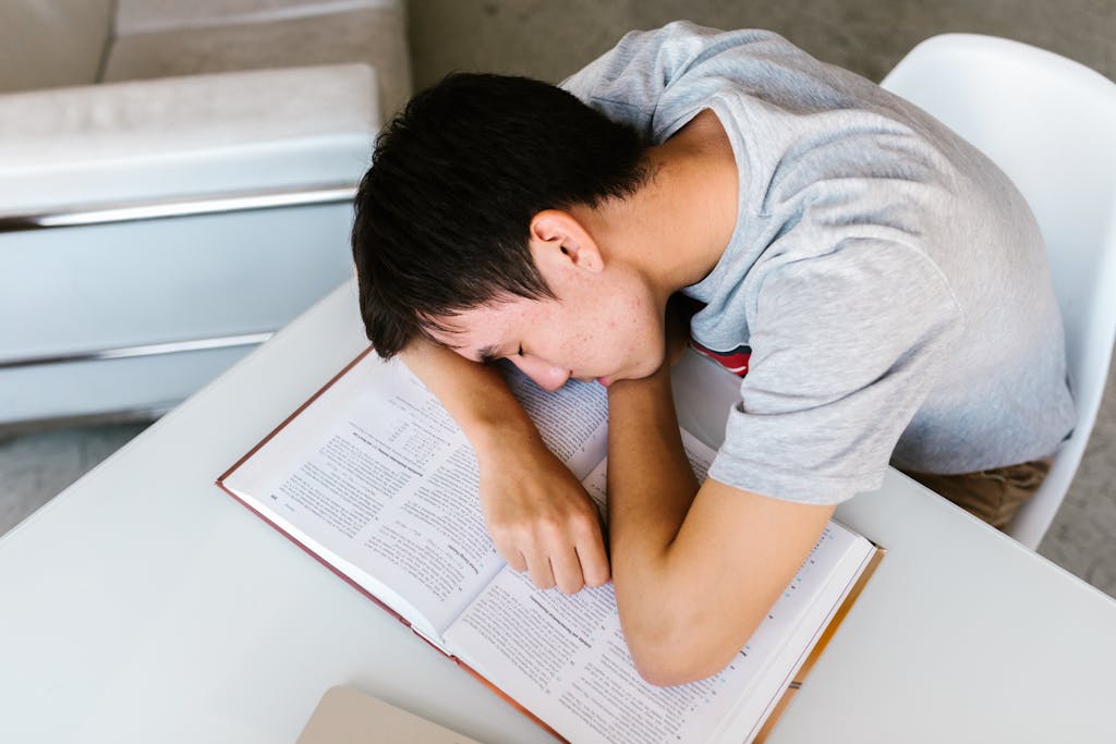 A young man sleeping on a white table with an open textbook, depicting study fatigue.