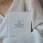 Close-up of a woman in a sweater holding The Five Minute Journal.